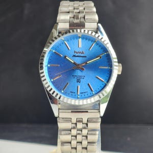 May include: A silver wristwatch with a blue face. The watch face displays the text "hmt Kohinoor" and "PARA SHOCK 17 JEWELS". It has a fluted bezel and a stainless steel strap.