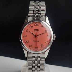 Puede incluir: Un reloj de pulsera vintage con una correa y caja de metal plateado. La esfera del reloj es de color salm&oacute;n con marcas y agujas negras. La esfera tiene texto en idioma extranjero y las palabras "PARA SHOCK 17 JEWELS".