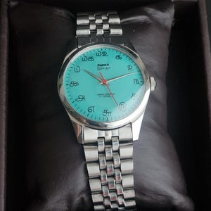 May include: A silver wristwatch with a turquoise face and a silver band. The watch face features black numerals and the text "hmt" and "PARA SHOCK 17 JEWELS". The watch is displayed on a dark brown cushion.
