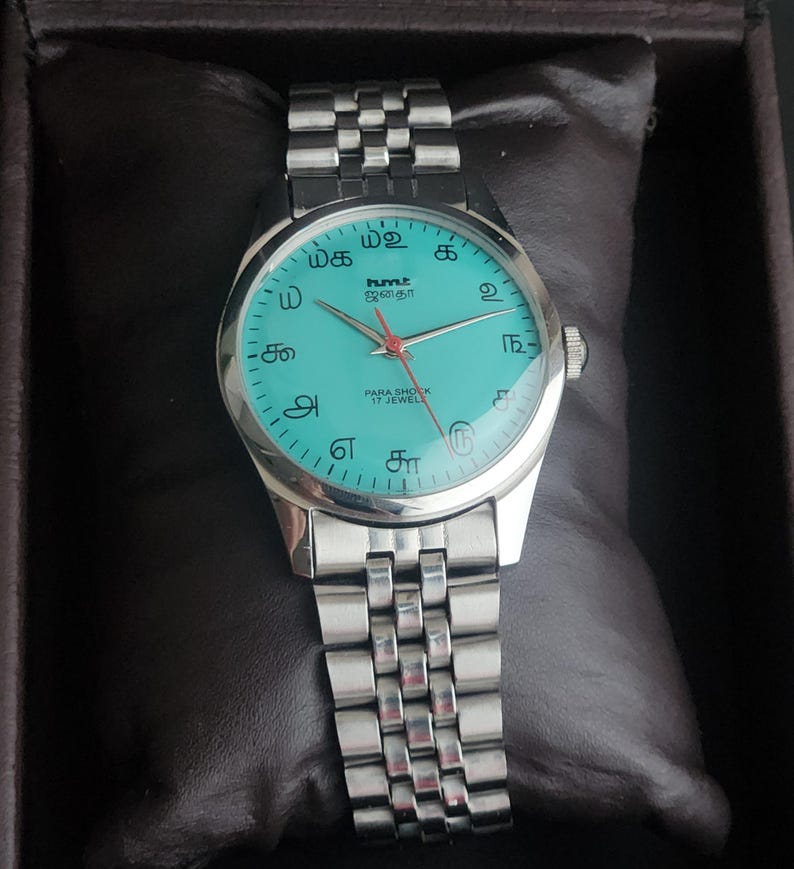May include: A silver wristwatch with a turquoise face and a silver band. The watch face features black numerals and the text "hmt" and "PARA SHOCK 17 JEWELS". The watch is displayed on a dark brown cushion.
