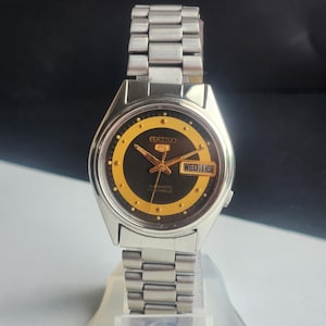 May include: A vintage Seiko 5 automatic wristwatch with a silver-tone stainless steel band. The watch face is black with a gold-colored ring and hands. The day and date are displayed at the 3 o'clock position. The watch is displayed on a clear stand.