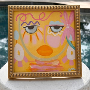 May include: A framed abstract painting of a face with pink, blue, and yellow colors. The face has large eyes, a pink nose, and a bright orange mouth. The background is a yellow color.