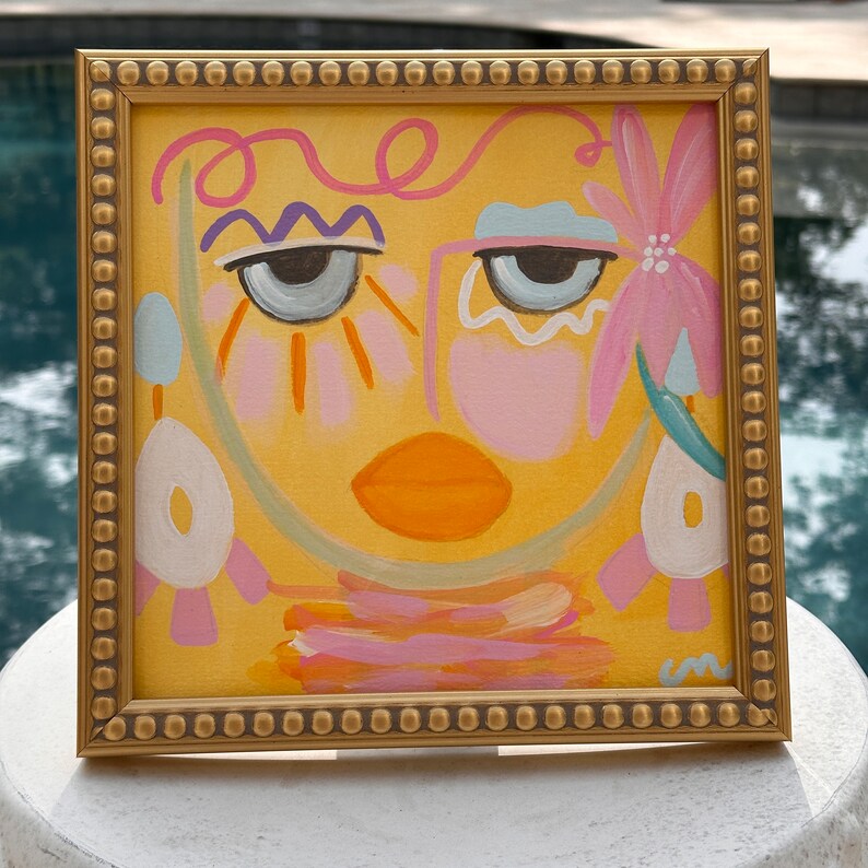 May include: A framed abstract painting of a face with pink, blue, and yellow colors. The face has large eyes, a pink nose, and a bright orange mouth. The background is a yellow color.
