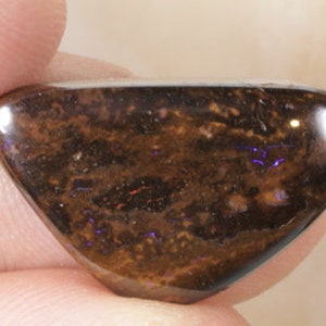 May include: A brown and black opal cabochon with iridescent purple and blue flashes. The opal is a teardrop shape and has a smooth, polished surface.