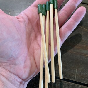 Può includere: Un gruppo di spiedini di bambù con punte verdi. Gli spiedini, tenuti in una mano, mostrano il loro colore naturale del legno e il verde vibrante delle estremità. Probabilmente per uso culinario.