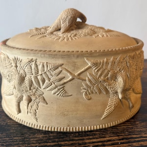 Może przedstawiać: Okrągłe, beżowe, nieszkliwione pudełko ceramiczne z pokrywką, ozdobione misternymi reliefami przedstawiającymi ptaki siedzące wśród liści paproci. Pokrywka zwieńczona jest rzeźbionym ptakiem, a podstawa otoczona ozdobnym koralikowym obrzeżem.