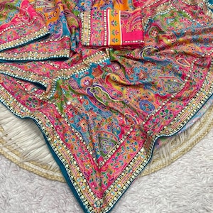 May include: A vibrant fabric textile featuring a multi-coloured paisley and floral pattern in shades of hot pink, turquoise, orange, and yellow. The material is adorned with small reflective mirror accents and a decorative border with gold trim.