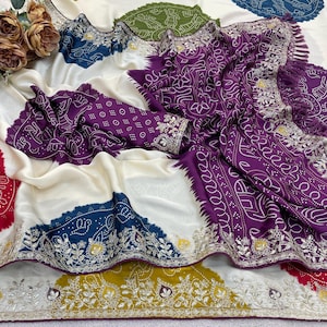 Puede incluir: Un sari blanco y morado con intrincados bordados y un diseño estampado. El sari presenta un borde y detalles morados, con adornos detallados. El diseño incluye una mezcla de colores, incluyendo azul, verde y amarillo.