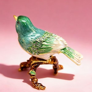 May include: A decorative bird figurine in shades of green and blue, perched on a gold-coloured branch. The bird's wings and body are adorned with small, sparkling embellishments. The figurine is set against a pink background.