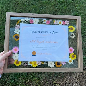 May include: A framed certificate with the text "Insert diploma here" and "A loyal customer." The frame is surrounded by colorful dried flowers, including sunflowers and daisies. The frame is held by a person.
