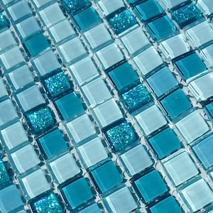 May include: A close-up of a blue and white mosaic tile sheet. The tiles are small squares with a glossy finish. Some of the blue tiles have a glittery texture.