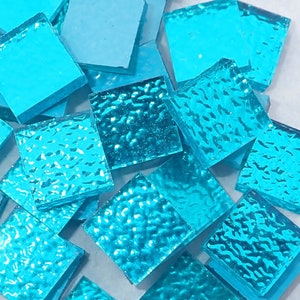 May include: A collection of small, square, turquoise glass tiles. Some tiles have a smooth surface, while others have a textured, rippled appearance. The tiles are scattered on a light-colored surface, creating a mosaic or craft supply theme.