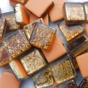 May include: A pile of small, square, orange and brown glass tiles with a gold metallic finish. The tiles are perfect for crafting and home decor projects.