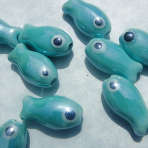 May include: A collection of teal blue glass fish beads with black eyes. The beads are all different sizes and shapes, and they have a glossy finish.