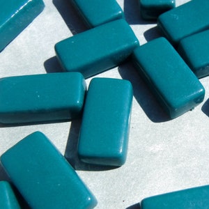 May include: Teal-colored rectangular glass beads scattered on a light surface. The beads have a glossy finish and a smooth, rounded shape. These beads are suitable for jewelry making and craft projects.