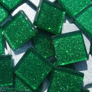May include: Close-up of square, emerald green glass tiles with glitter embedded within. The tiles are scattered on a light gray, textured surface. The tiles are likely for mosaic art or decorative projects.
