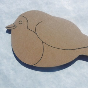 May include: A wooden bird cutout in a light brown color. The bird has a simple design with black outline details. The bird is lying on a white surface. The bird's beak is pointing to the left.