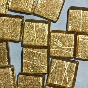 May include: A collection of small, square, gold-colored glass tiles. Each tile has a textured surface with subtle linear patterns. The tiles are scattered on a light blue surface, creating a shimmering effect. These tiles could be used for mosaics or decorative projects.