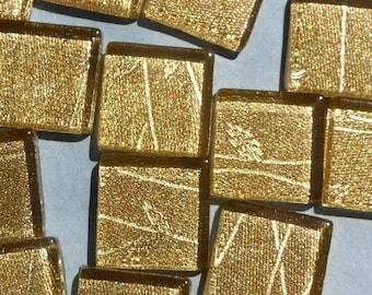 May include: A collection of small, square, gold-colored glass tiles. Each tile has a textured surface with subtle linear patterns. The tiles are scattered on a light blue surface, creating a shimmering effect. These tiles could be used for mosaics or decorative projects.