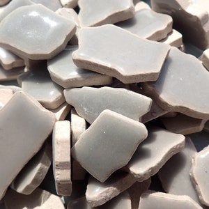 May include: A pile of irregularly shaped, light gray ceramic tiles with a glossy finish. The tiles have a slightly rough, unglazed edge, and vary in size and shape. The overall effect is a collection of broken or fragmented pieces.