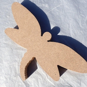 May include: A wooden dragonfly cutout with a natural, unfinished appearance. The wings are spread wide, and the body is detailed with a small head and segmented tail. The cutout is likely for crafts or decoration.