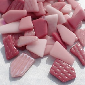 May include: A pile of pink and red glass mosaic tiles with a textured surface. The tiles are various shapes and sizes.
