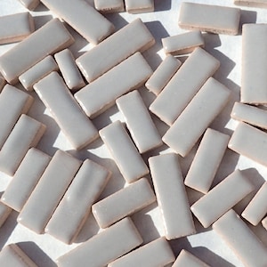 May include: A collection of small, rectangular ceramic tiles in a light gray color. The tiles are arranged in a random pattern on a white surface.