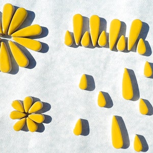 May include: Bright yellow, petal-shaped ceramic tiles arranged in floral and abstract patterns. The tiles are on a white surface, with some arranged in the shape of flowers, and others scattered. The tiles have a glossy finish.