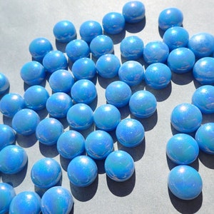 Puede incluir: Un conjunto de canicas de vidrio azul iridiscente dispersas sobre una superficie gris. Las canicas tienen un acabado brillante y reflejan la luz, creando un efecto brillante. Estas canicas decorativas son adecuadas para juegos o manualidades.