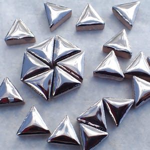 May include: A collection of shiny, silver-colored triangular tiles. The tiles have a reflective surface and are arranged in a scattered pattern, with some forming a hexagonal shape. The edges of the tiles are a light brown color.