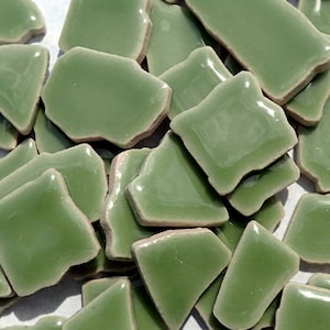 May include: A pile of irregularly shaped, glossy green ceramic tiles. The tiles have a smooth, reflective surface and a slightly uneven edge. The color is a muted, earthy green.