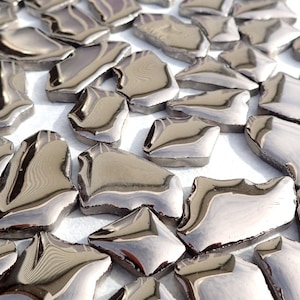 May include: A collection of irregularly shaped, silver-colored mosaic tiles. Each tile has a reflective, metallic surface and a dark border. The tiles are scattered across a white surface, creating a textured appearance.