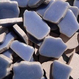 May include: A collection of irregular-shaped, light blue ceramic tiles with rough, off-white edges. The tiles are scattered, creating a textured surface. The color palette is soft, with a focus on the cool blue and neutral tones. These tiles are suitable for mosaic projects.