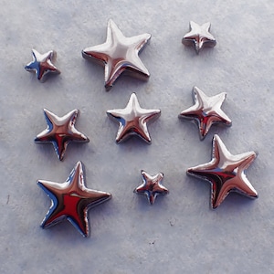 May include: A collection of shiny, silver-colored star-shaped embellishments of varying sizes. The stars have a reflective surface, creating highlights and shadows. The arrangement of the stars is scattered across a light-colored surface.