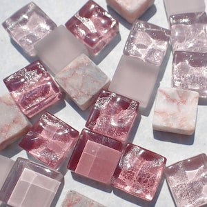 May include: A collection of square mosaic tiles in various shades of pink and white. Some tiles are solid, while others have a marbled or textured appearance. The tiles are scattered, creating a visually appealing arrangement.