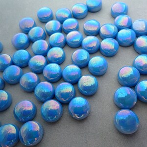 Puede incluir: Una colecci&oacute;n de gemas de vidrio azul iridiscente. Cada gema es un disco redondeado de fondo plano, que refleja un espectro de colores. Las gemas est&aacute;n dispersas sobre una superficie neutra, creando un efecto brillante.