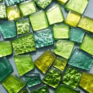 May include: A selection of square glass tiles in various shades of green and yellow. The tiles display different textures and colours, from deep emerald to bright lime. Some tiles have a textured surface, while others are smooth. The tiles are scattered on a light grey surface.