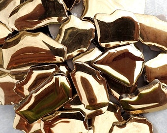 May include: A collection of irregularly shaped, gold-colored ceramic pieces. The pieces have a reflective, metallic finish, and are scattered across a surface. The shapes are reminiscent of broken fragments.
