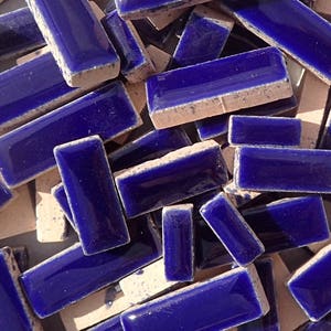 May include: A close-up of numerous rectangular, deep blue ceramic tiles. Each tile has a glossy surface and a rough, beige underside. These tiles are suitable for mosaic work or decorative purposes, and are approximately 5 cm long.