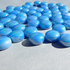 Puede incluir: Una colecci&oacute;n de guijarros de vidrio azul iridiscente. Las piedras lisas, de forma ovalada, tienen un acabado brillante y reflejan la luz, creando un efecto brillante. Los guijarros est&aacute;n dispersos sobre una superficie gris claro.