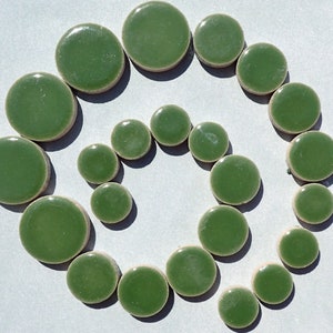 May include: A collection of green, circular ceramic tiles arranged in a spiral pattern. The tiles vary in size, with a glossy finish and a light brown edge. The background is a light gray.