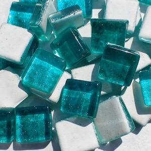 May include: Close-up of a collection of square glass tiles in shades of teal and white. The tiles are scattered, with some translucent teal tiles reflecting light. The white tiles have a textured, opaque appearance. Ideal for mosaic projects.
