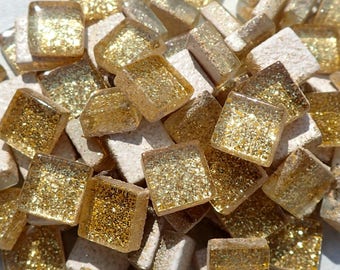 May include: Close-up of a collection of square and rectangular mosaic tiles. The tiles are a mix of gold glitter and off-white, creating a shimmering effect. The tiles are likely for decorative purposes, such as for a backsplash or other craft projects.