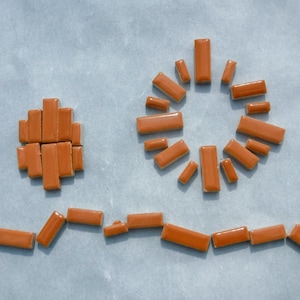 May include: A collection of small, rectangular, brown ceramic tiles arranged in the shape of a sun, a mountain range, and a circle.