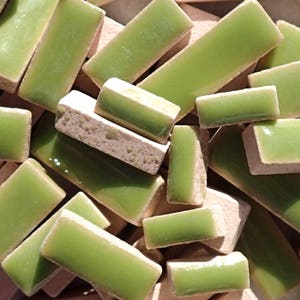 May include: A collection of rectangular green tiles with a glossy surface. The tiles are arranged in a pile, some showing a beige, porous underside. The image presents a textured, colourful display of building materials.