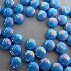 Puede incluir: Un conjunto de canicas de vidrio azul iridiscente. Cada canica tiene forma redondeada y un acabado brillante, reflejando un espectro de colores. Las canicas est&aacute;n dispersas sobre una superficie gris, creando una exhibici&oacute;n visualmente atractiva.