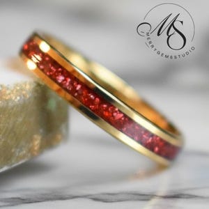 May include: A gold-toned ring featuring a central inlay of crushed red material with a sparkling texture. The band has a smooth, polished finish on the edges. A circular logo with the text MERRY GEMS STUDIO is visible in the upper right corner.