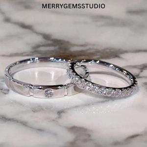 Op de afbeelding: Twee zilveren ringen. Eén ring heeft een enkele, ronde diamant in het midden, gezet in een rechthoekig ontwerp. De andere ring is volledig ingelegd met kleine, sprankelende diamanten. De ringen worden gepresenteerd op een marmeren oppervlak.