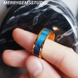 May include: A gold-colored ring with a blue wood inlay. The ring is held up by a finger. The text "MERRYGEMSSTUDIO" is in the top left corner.