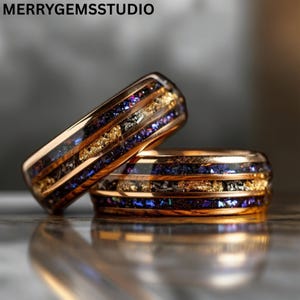Puede incluir: Dos anillos de oro rosa con un diseño único. Cada anillo presenta una banda de copos de oro e incrustaciones de color azul oscuro y púrpura. Los anillos se muestran sobre una superficie reflectante. El texto "MERRYGEMSSTUDIO" está en la parte superior.